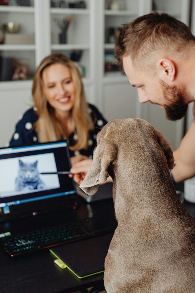 A dog and two adults work together in a home office, looking at a laptop screen.